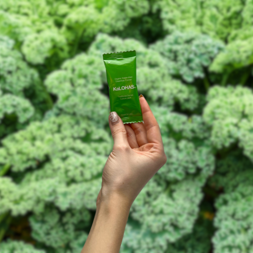 A hand holding a KaLOHAS sachet in front of fresh kale