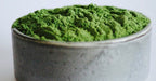 A close-up of bright green kale powder, piled in a small round gray ceramic bowl against a plain light background.