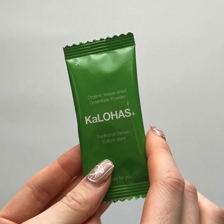 A hand holding a green foil packet of KaLOHAS+ Organic freeze-dried GreenKale Powder against a plain light gray background.